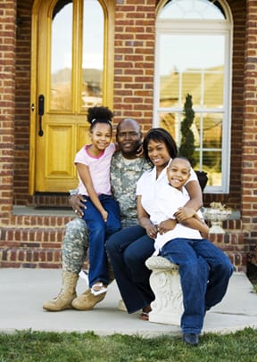 A family sitting in front of a home. Father is wearing military fatigues. 