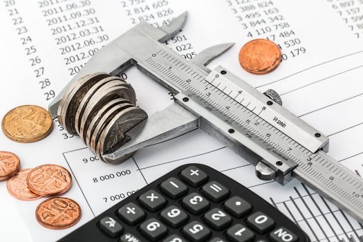 Calipers holding coins beside a calculator, both laying on a financial spreadsheet.