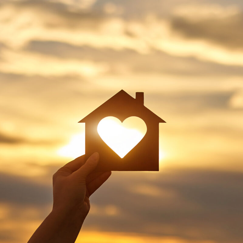 Woman hand holds wooden house in the form of heart against the sun. Solar energy. Children dreams. International day of families. Home protection insurance concept. Planning to buy property. A symbol for ecology.