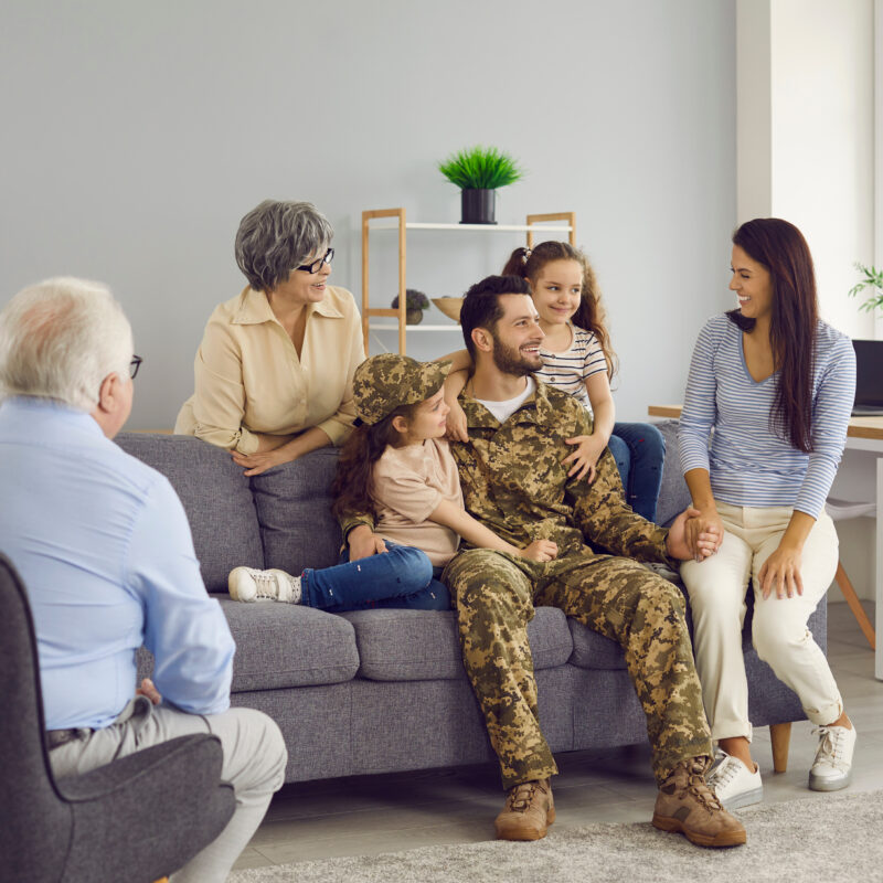 Returning to the family. Happy soldier man is talking with his family after returning from military service sitting in the living room at home. Older parents, wife and children all rejoice together.