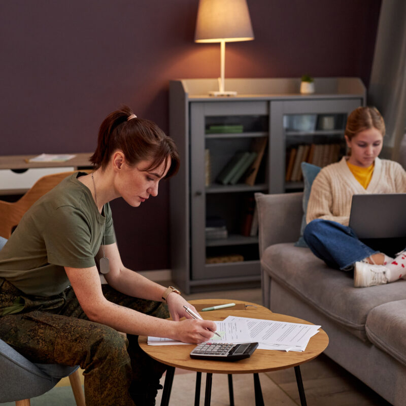 Side view portrait of young military woman working with documents at home, daughter in background, copy space