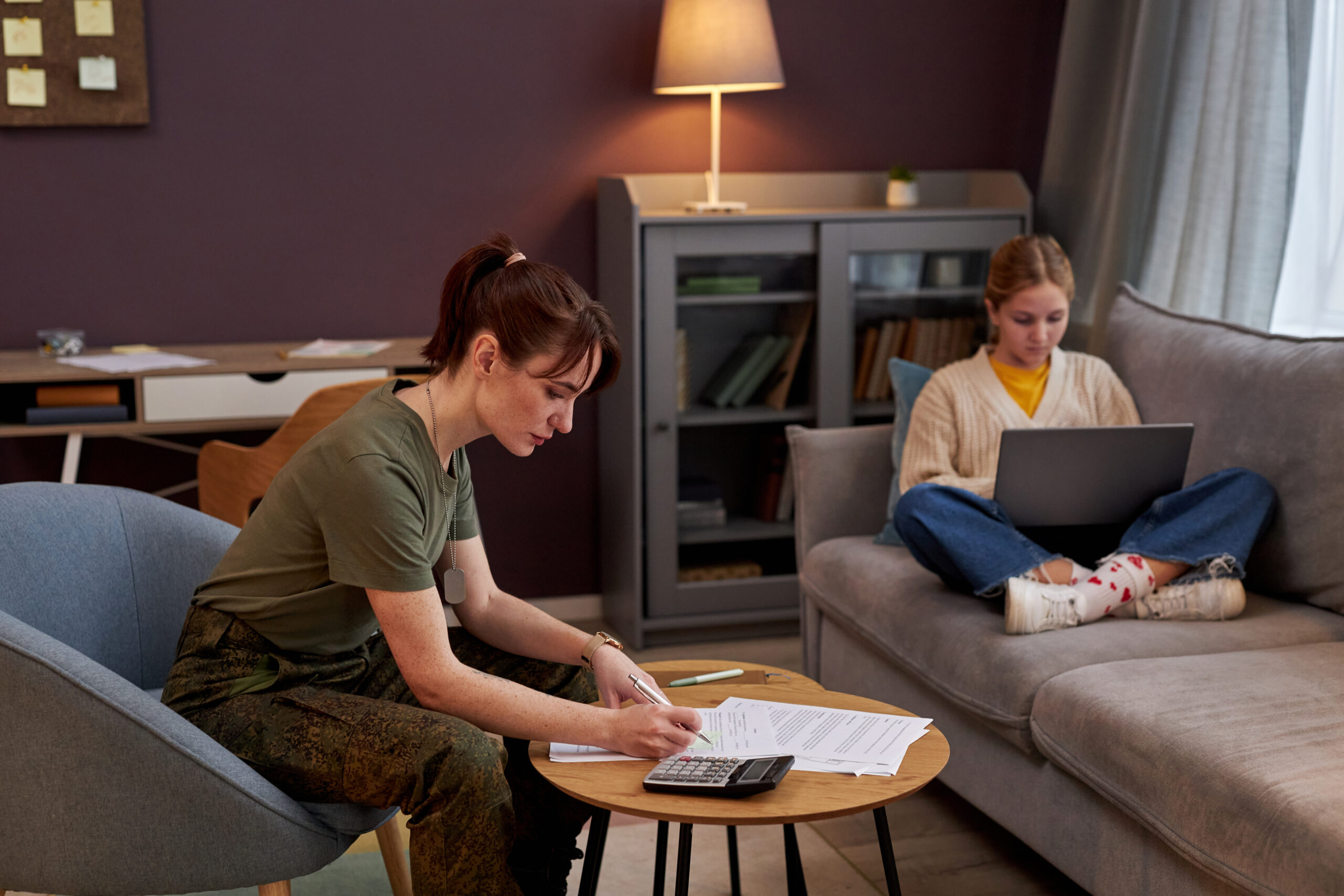 Side view portrait of young military woman working with documents at home, daughter in background, copy space