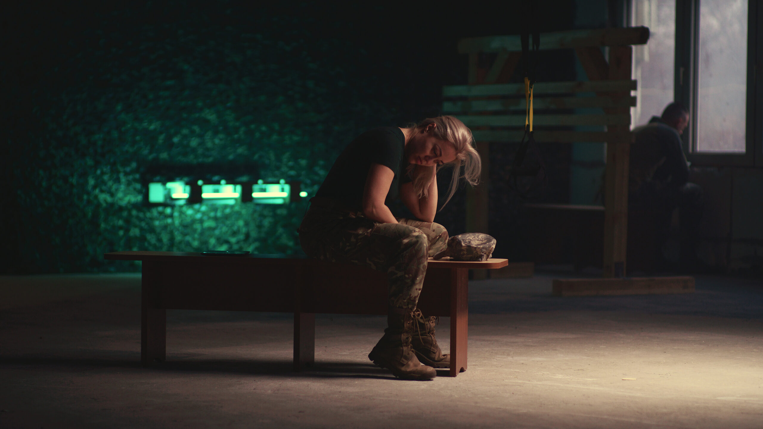 View of tired military woman taking off cap and touching hair while sitting on bench and relaxing in dark gym