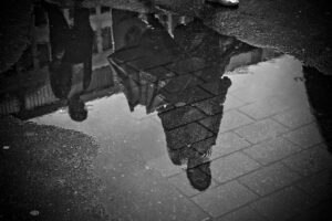 In this back and white photo a two people and the buildings behind them are reflected in a rain puddle on the street.