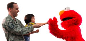 Service member holding young child to high-five Elmo