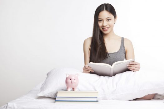 Young woman sitting on bed with books and piggy bank
