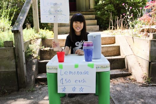 A young girl behind a lemonade stand, holding a sign that says "lemonade 50 cents each."