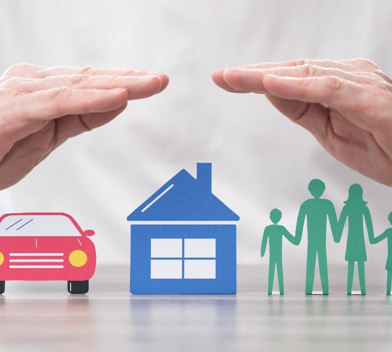 Man's hands hovering protectively over paper cutouts of a house, car, and family lined up on a table.