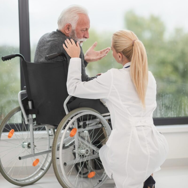 Elderly gentleman in a wheel chair with a healthcare worker kneeling to talk to him