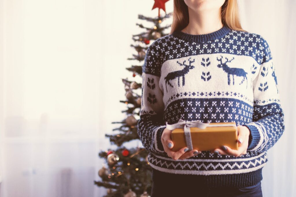 Woman wearing blue holiday sweater holding wrapped gift in front of Christmas tree.