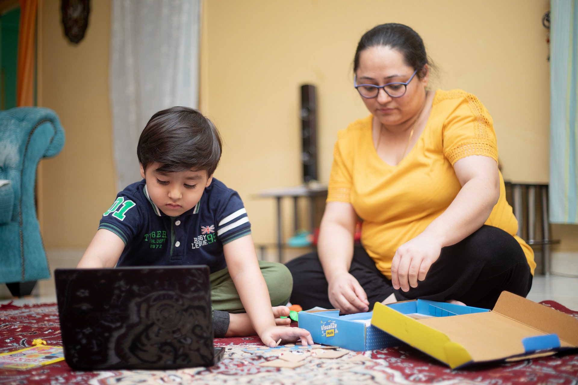 A young boy and a female caregiver sit on the floor with a laptop for online learning