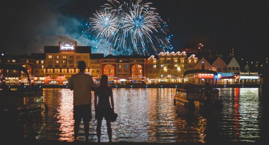 A man and woman in foreground facing away from camera looking over a night sky and river with skyline in the background and fireworks shooting up over the buildings.