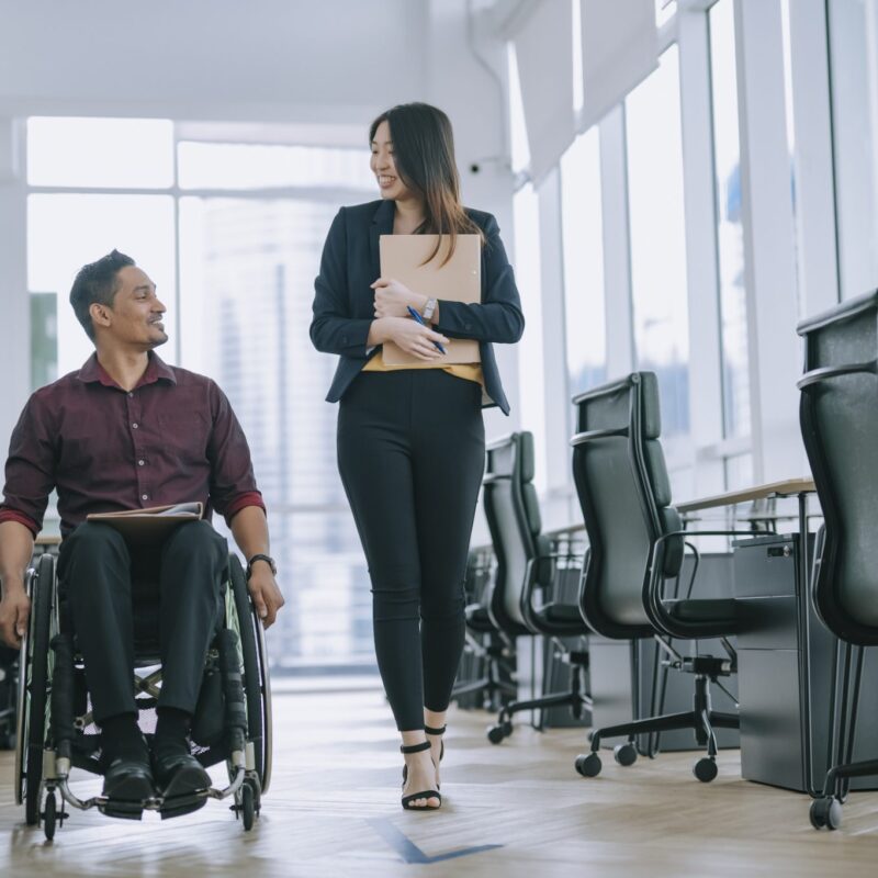 Indian white collar male worker in wheelchair having cheerful discussion conversation with his female asain chinese colleague coworking at walkway corridor