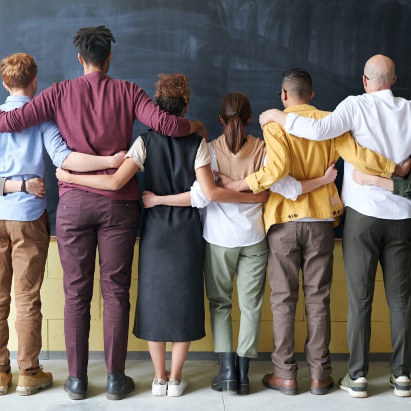 Multicultural group of people facing a chalkboard with arms interlocked.