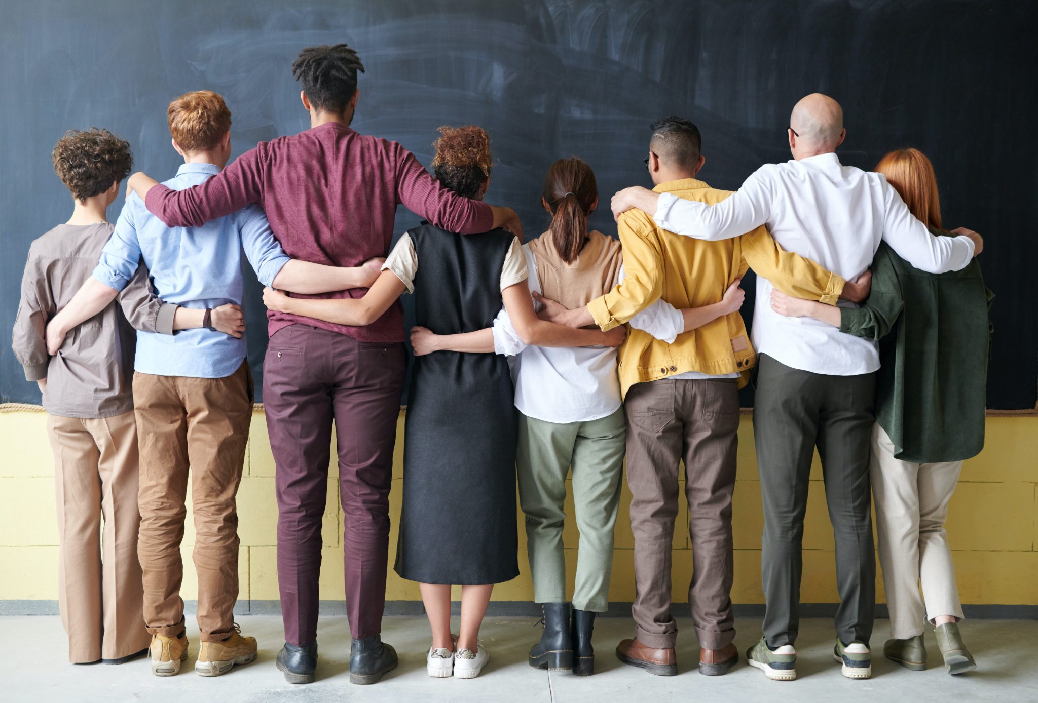 Multicultural group of people facing a chalkboard with arms interlocked.