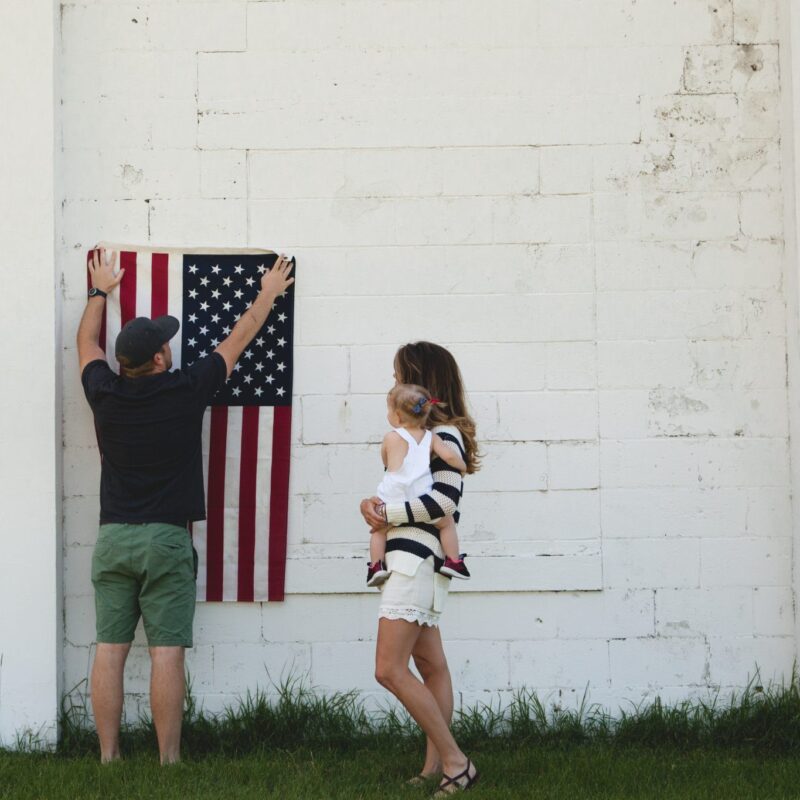 Photo of woman holding baby while man puts up American flag on building