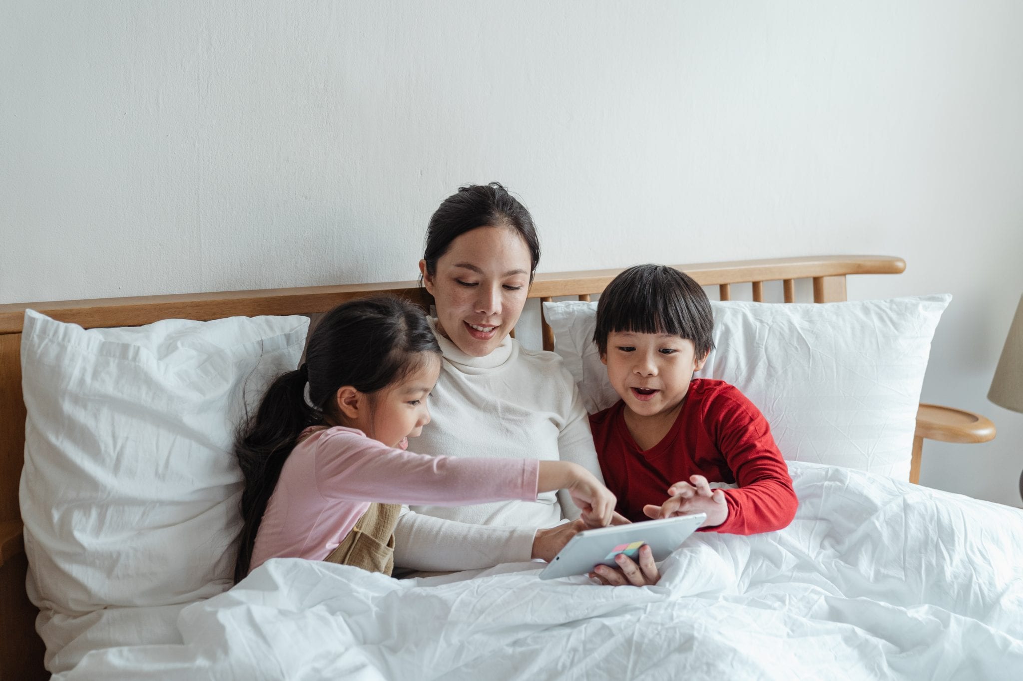 photo of woman and kids sitting on bed while using tablet computer