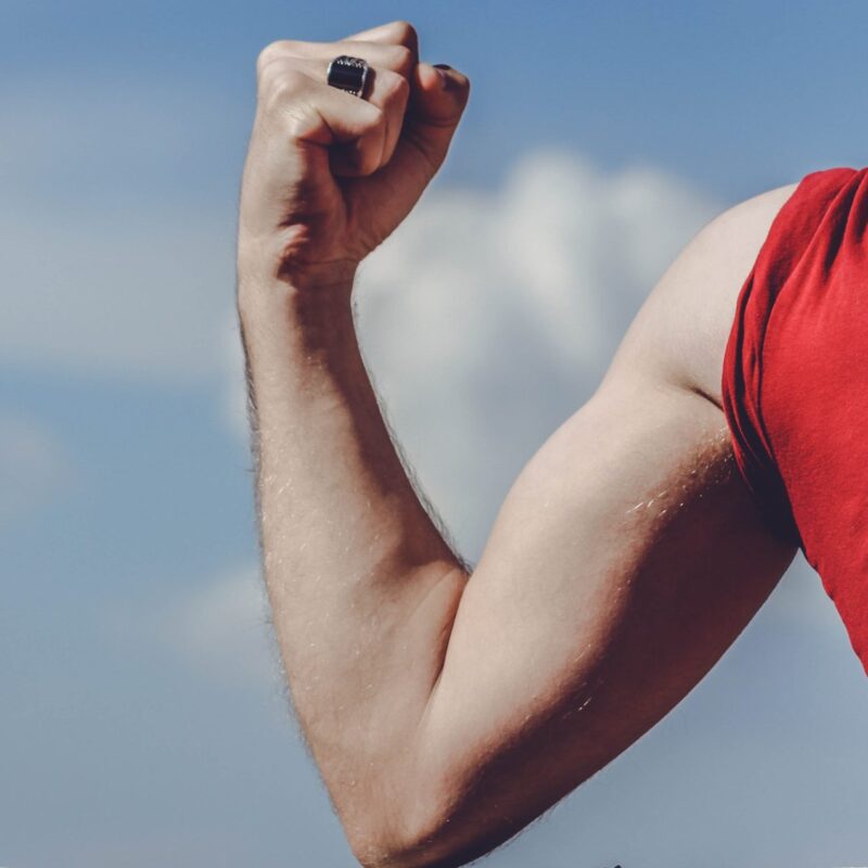 Person in red shirt flexing bicep muscle against blue sky with clouds