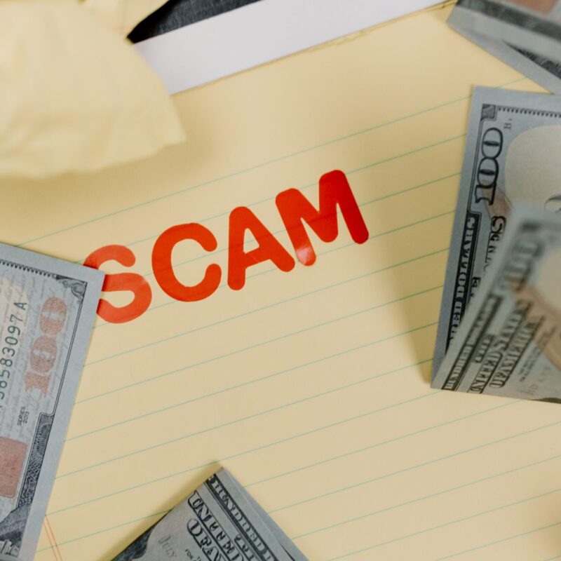 The word "scam" on a yellow background with cash around it.