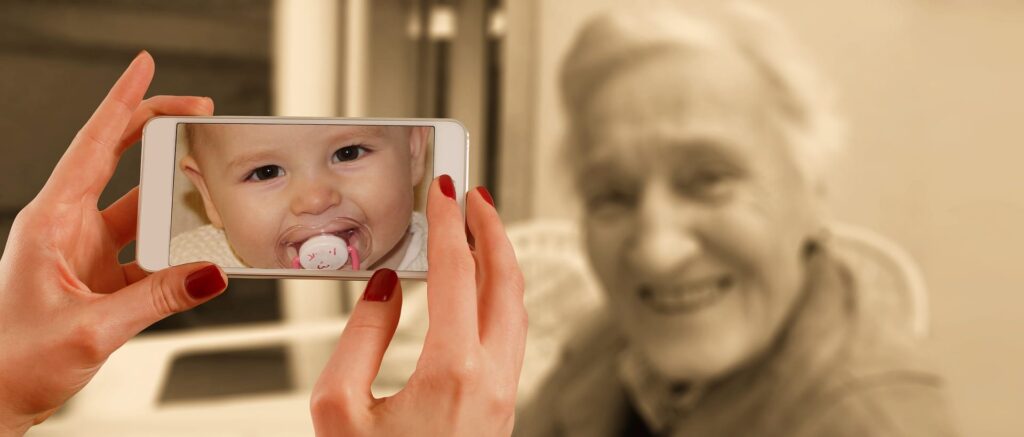 A photo of a woman's hands holding a smart phone with a picture of an infant on it, in the background a faded photo of an elderly woman.