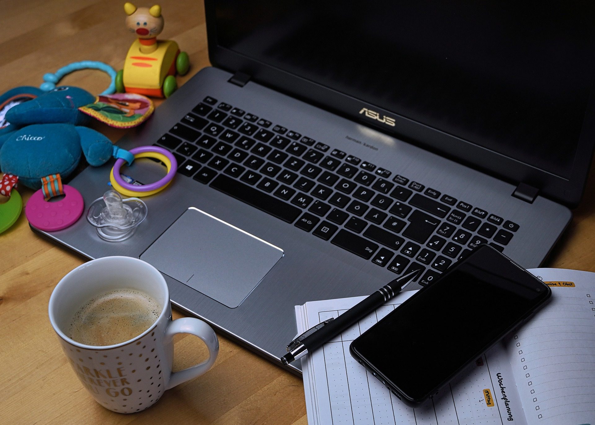 Image of laptop with cell phone, coffee, baby toys, planner to depict the struggle of balancing online learning