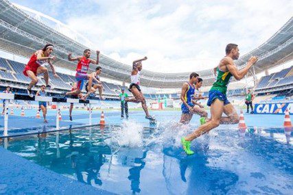 Male track athletes jumping hurdles into water.