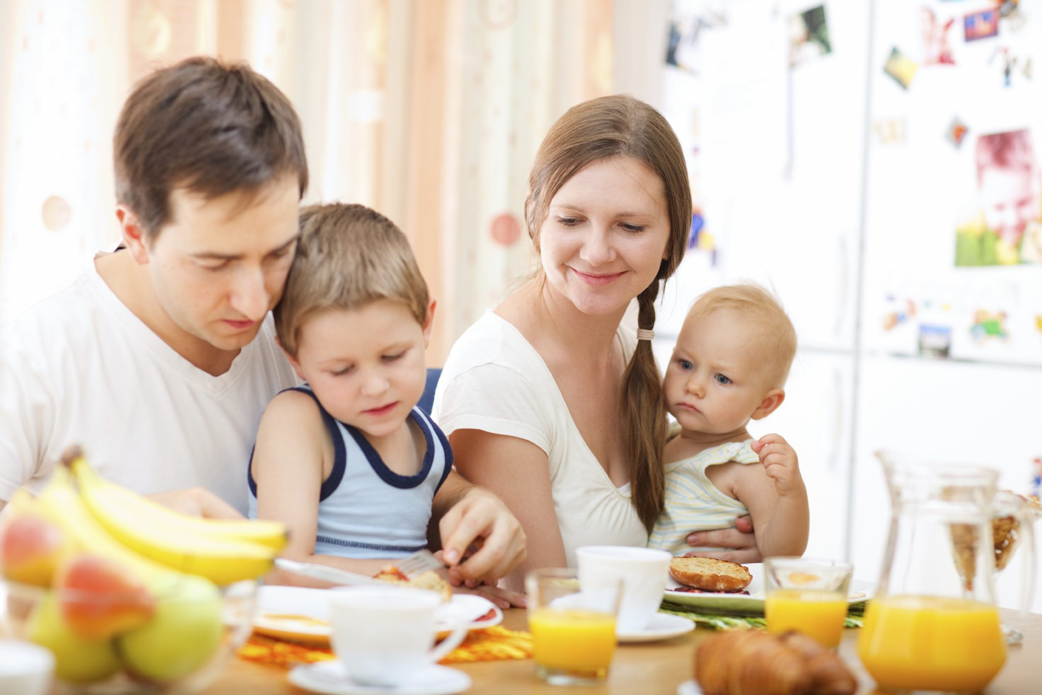 Family with small children eating breakfast. Dad holding toddler; mother holding baby.