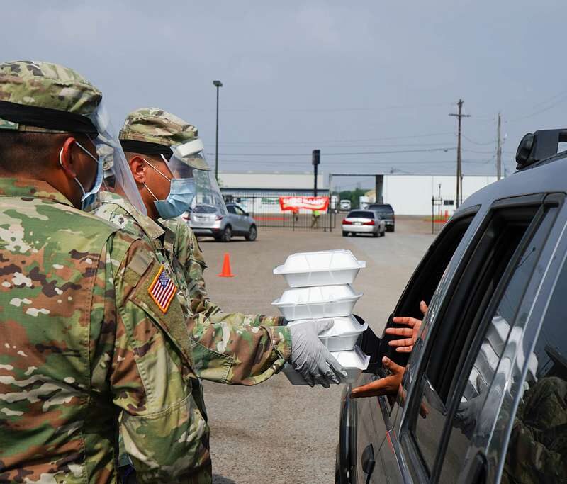 Two soldiers hand a stack of white styrofoam foos containers to a person reaching out the window of a dark colored SUV.