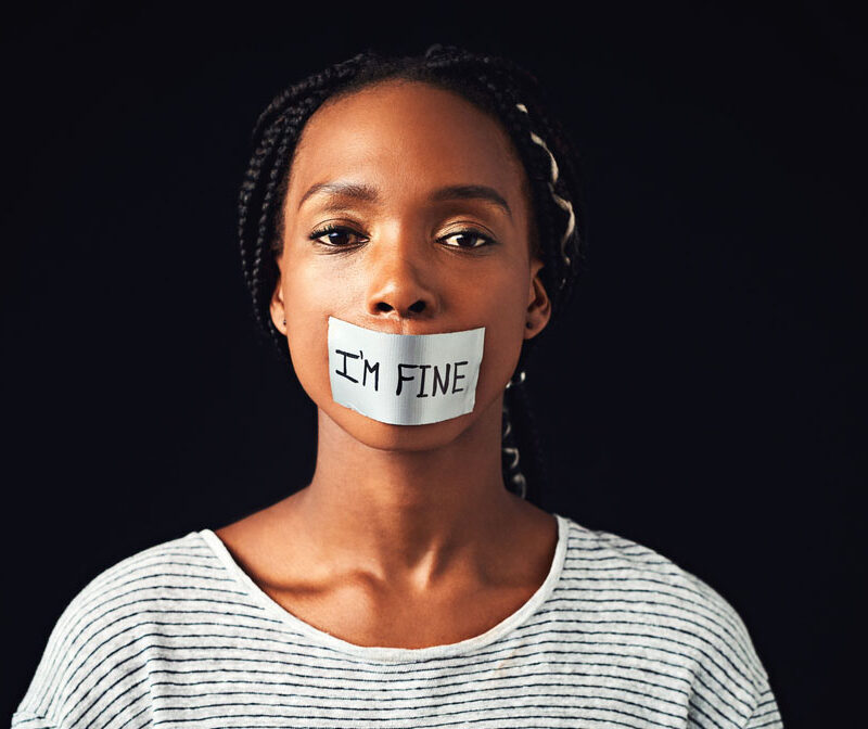 Studio shot of a young woman with a label saying “I’m fine” covering her mouth against a black background