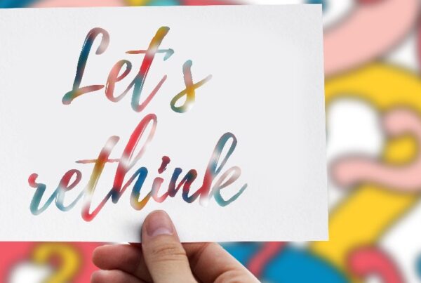 A person's hand holds a white card with the words "Let's Rethink" cut out of it. A background of multicolored question marks can be seen through the cutout.