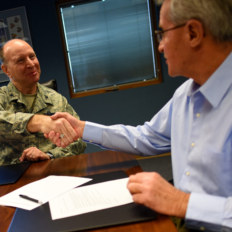 A man in U.S. Air Force uniform shakes hands across a table with a man in a blue button-up shirt.