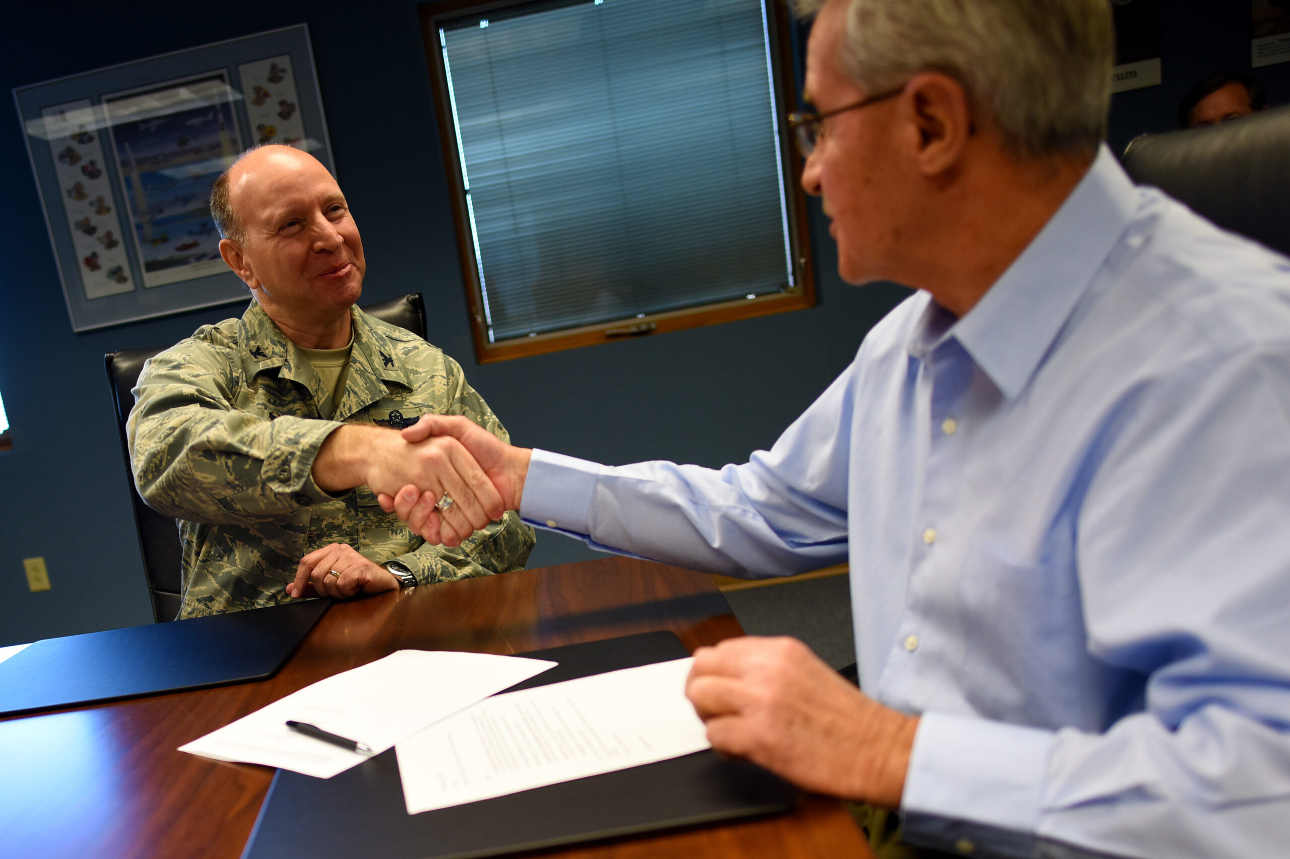 A man in U.S. Air Force uniform shakes hands across a table with a man in a blue button-up shirt.