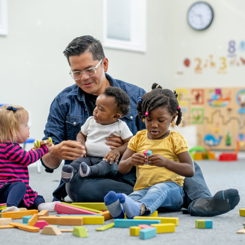 Childcare professional plays with three small children while sitting on the floor.