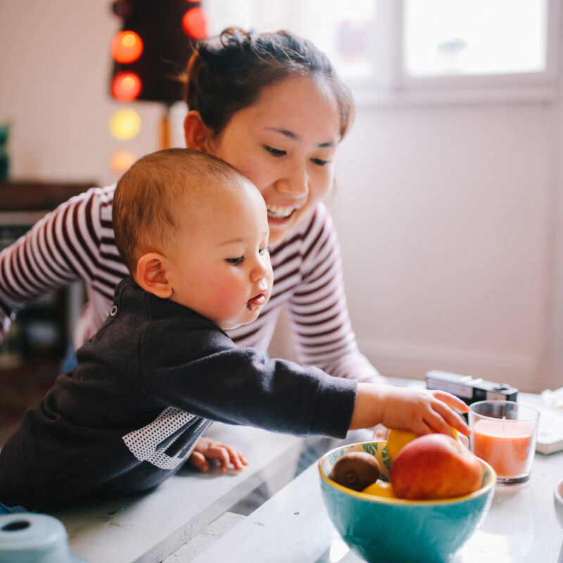 Brightly lit image of a young Thai mom with her little baby boy. They're spending time together at home, playing with toys and having some snacks. Asian family lifestyle concepts, shot in Europe.