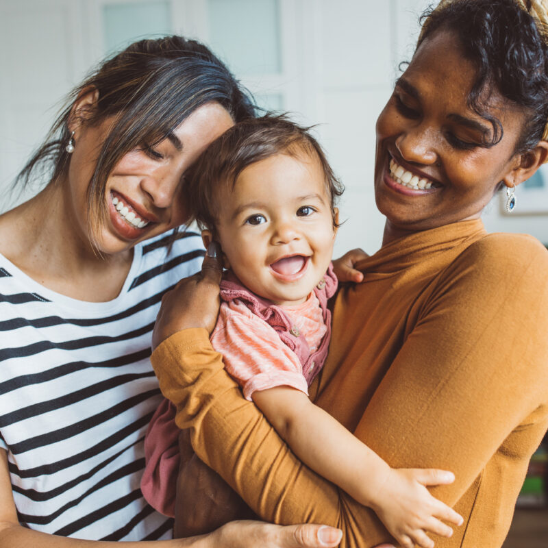Two smiling black or multiracial women holding a happy baby while all embrace