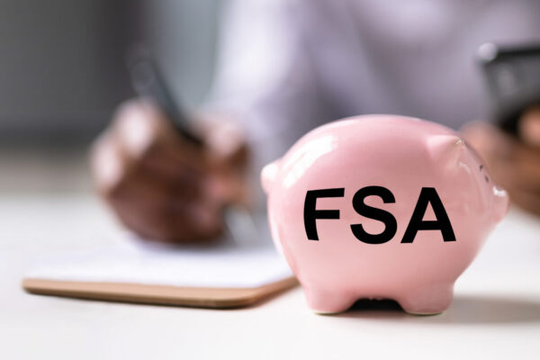 FSA written on a pink piggy bank. Background includes person writing on a note pad and holding a phone.