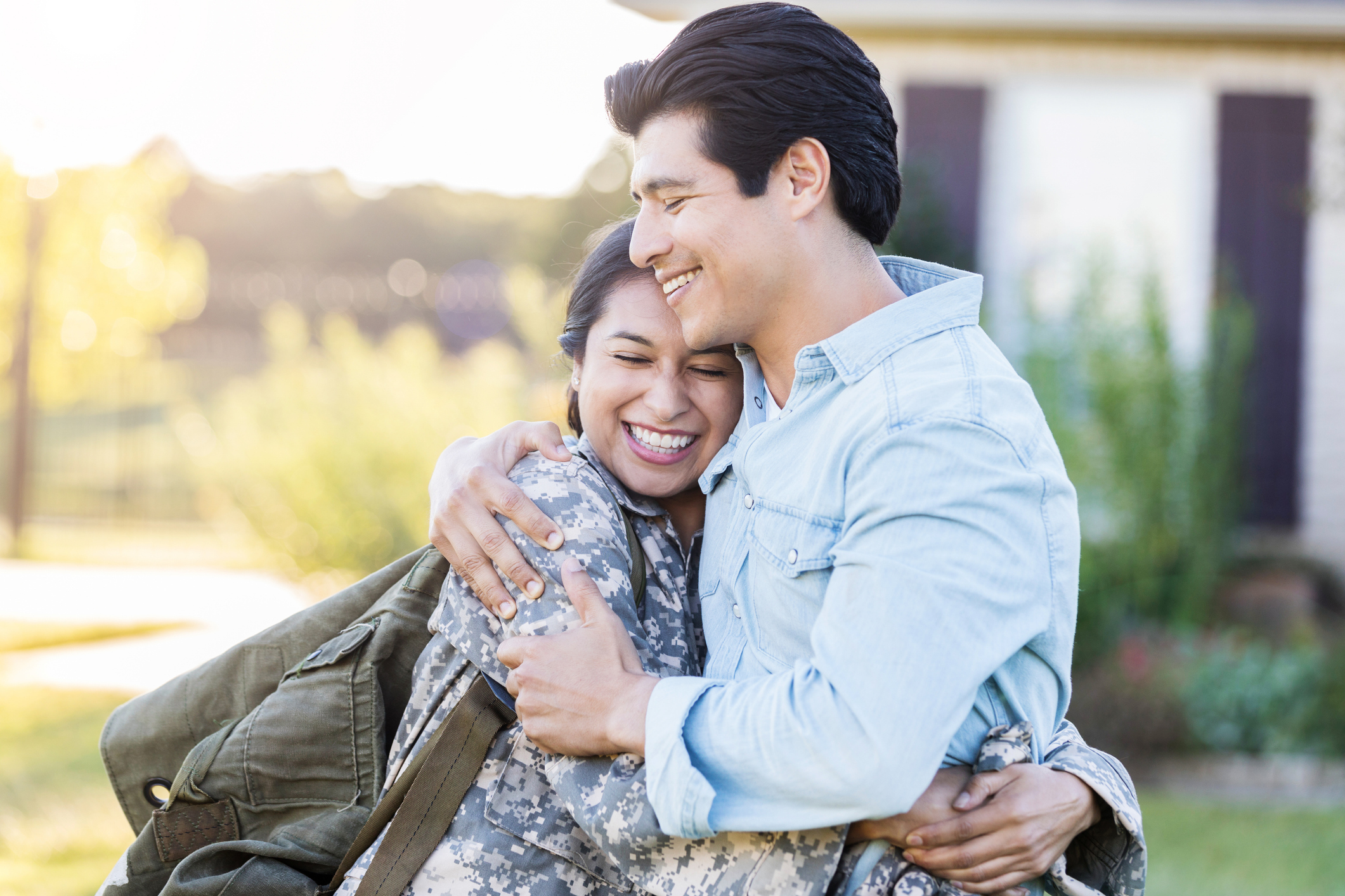 Young, Hispanic female in uniform hugs her husband tightly, after she has just arrived home from duty. She still holds her bag on her shoulder, while they stand outside their home.
