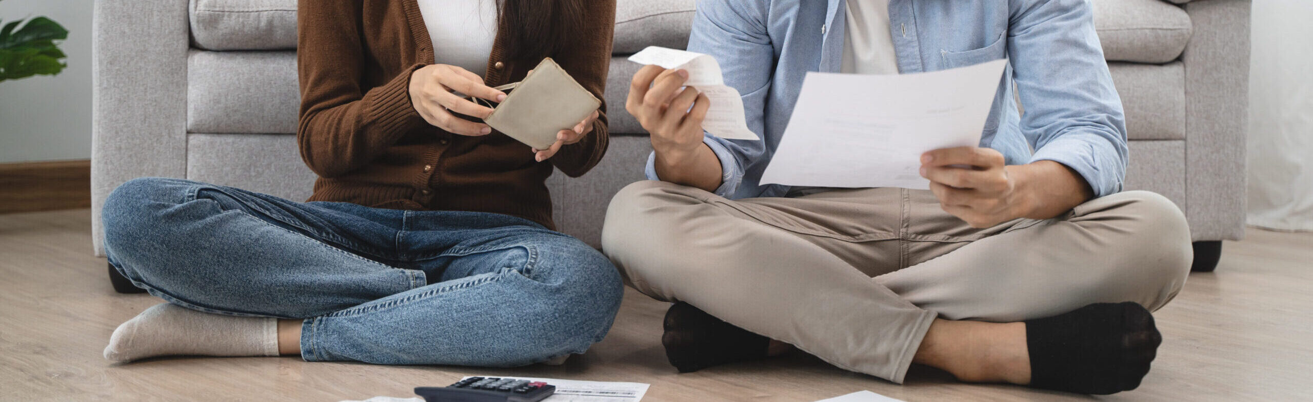 couple sitting on the floor with crossed legs, holding receipts and bills. Calculator on the floor.