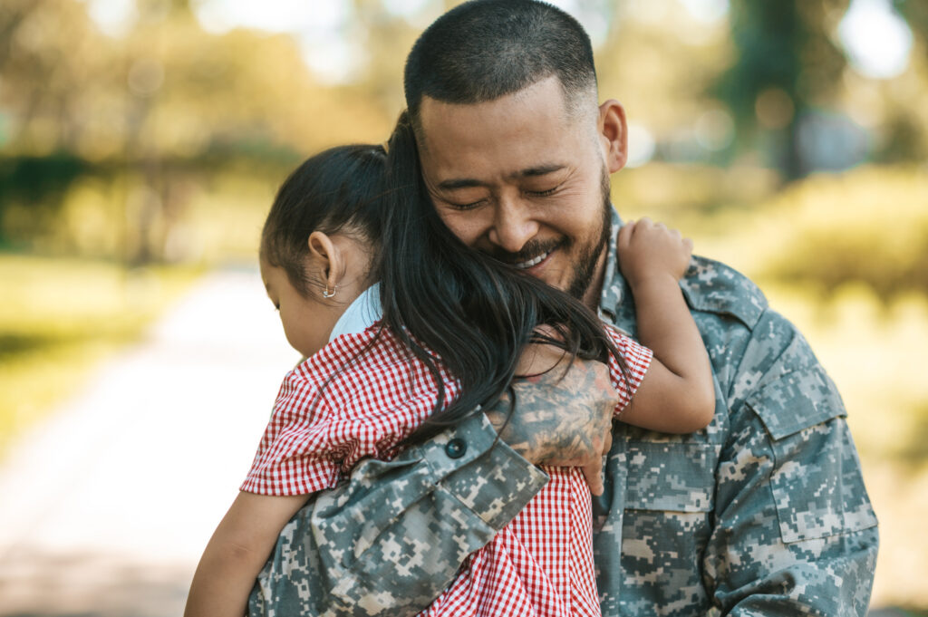 Father wearing military fatigues hugs young girl in red and white checked dress.