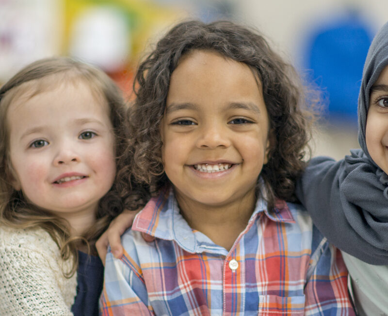 Two girls and a boy are indoors in their preschool classroom. They are smiling at the camera while sitting on the carpet and embracing.