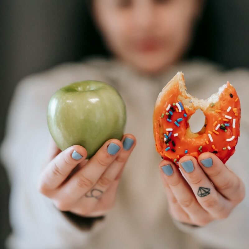 Child holding a green apple in right hand and sprinkle-covered donut in left hand.