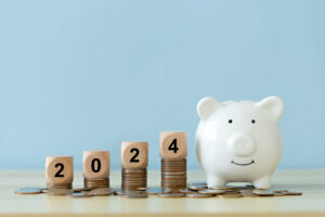 Text 2024 on a wooden cube on coins stacked with a white piggy bank and blue background.