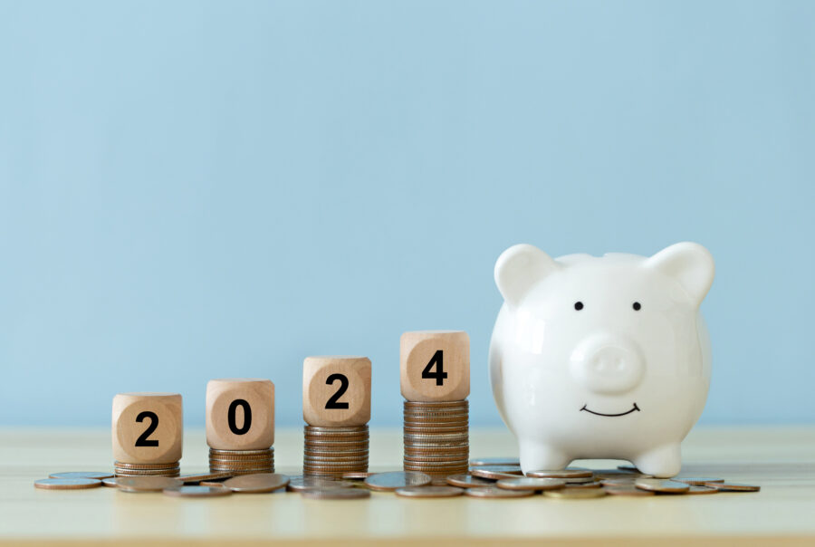 Text 2024 on a wooden cube on coins stacked with a white piggy bank and blue background.