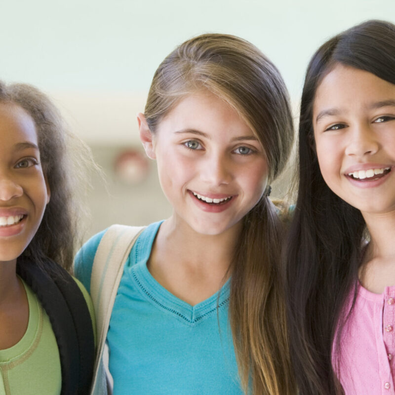 A group of female elementary school friends smiling in the hallway with their arms around each other