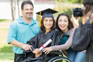 Happy Hispanic female college graduate in a wheelchair smiles for a photo with her parents. The graduate is sitting in a wheelchair. A college friend is taking a photo of the family.