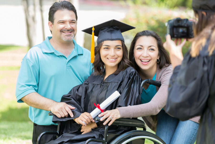 Happy Hispanic female college graduate in a wheelchair smiles for a photo with her parents. The graduate is sitting in a wheelchair. A college friend is taking a photo of the family.