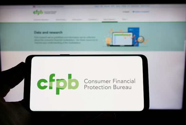 Person holding smartphone with logo of US Consumer Financial Protection Bureau (CFPB) on screen in front of website. Focus on phone display.