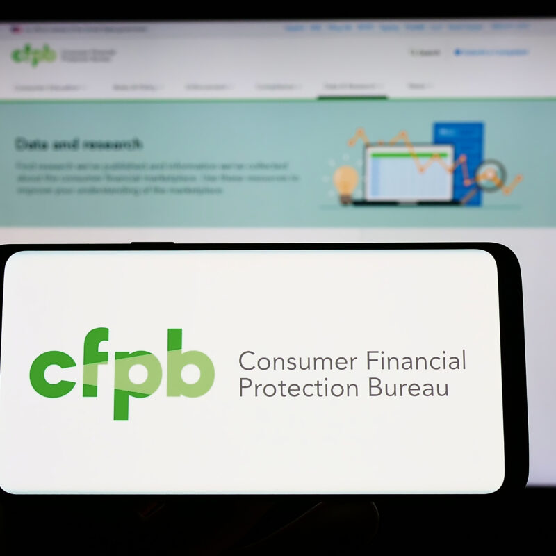 Person holding smartphone with logo of US Consumer Financial Protection Bureau (CFPB) on screen in front of website. Focus on phone display.