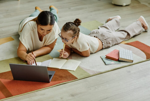 Mother helping girl with down syndrome studying at home while lying on floor together, non traditional education