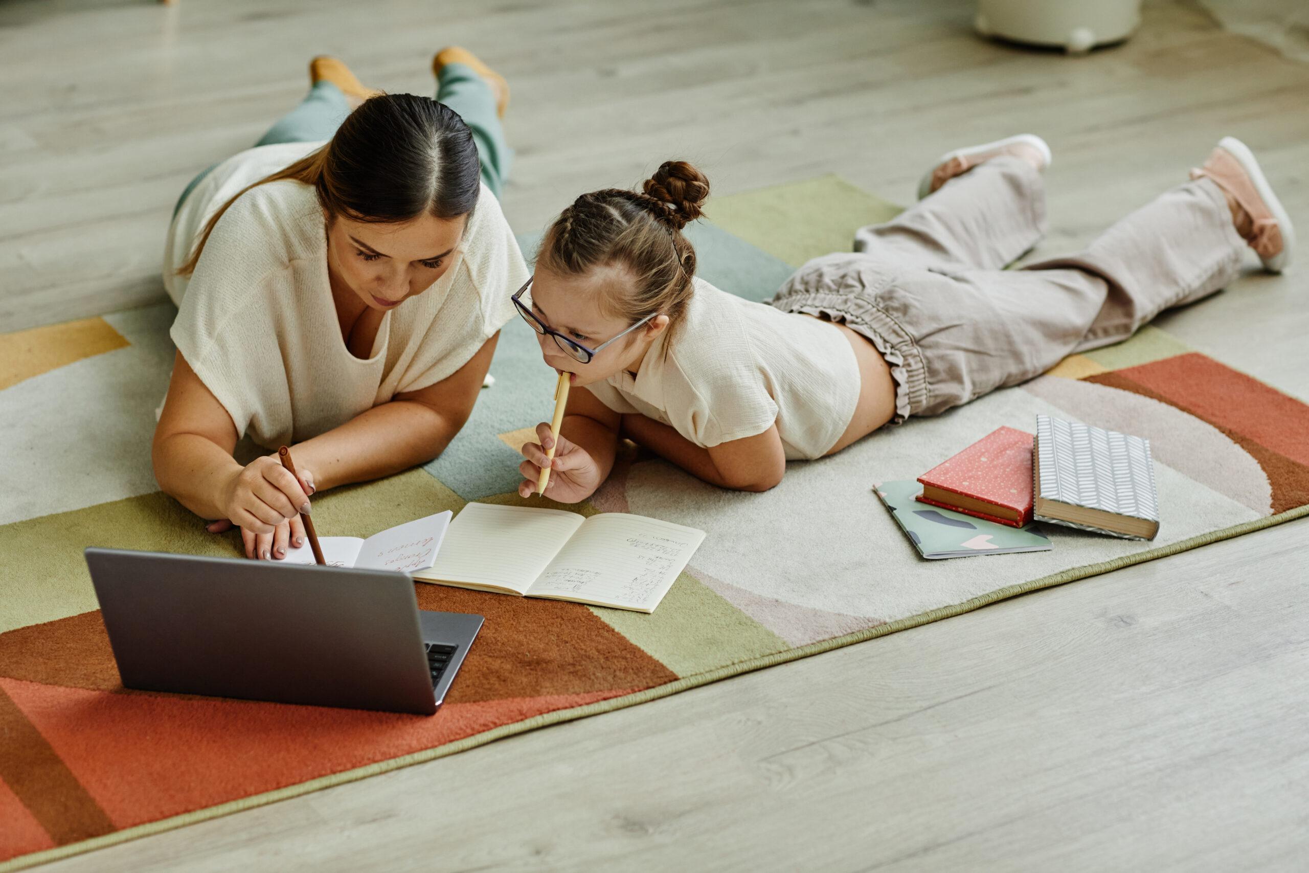 Mother helping girl with down syndrome studying at home while lying on floor together, non traditional education